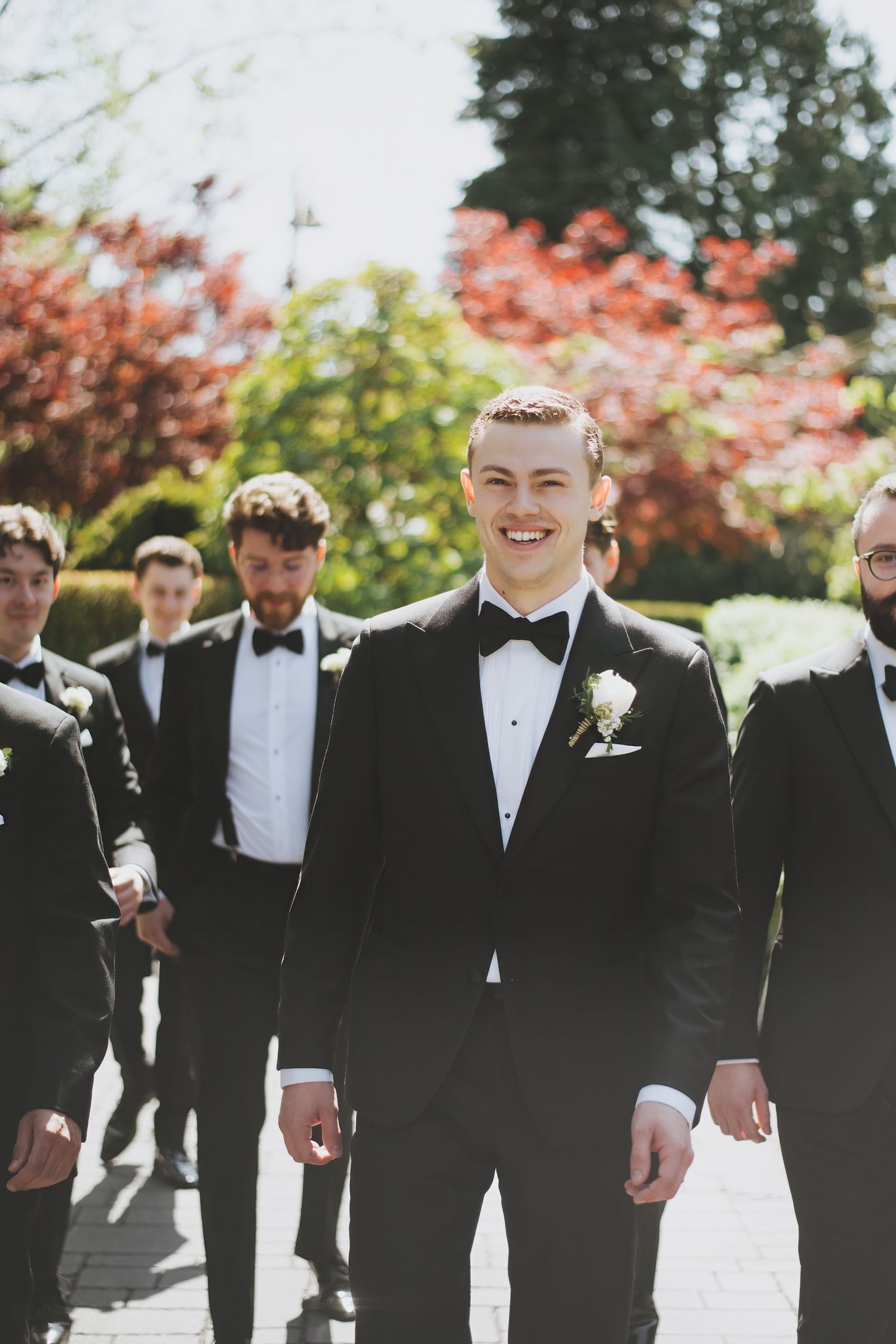 Groomsmen walking together outdoors in black tuxedos and bowties on sunny day with spring foliage in background.