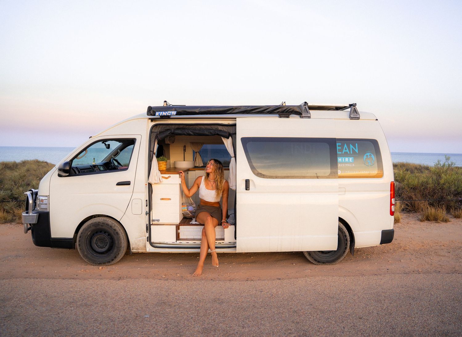A white campervan with open sliding door parked against an ocean backdrop at sunset.