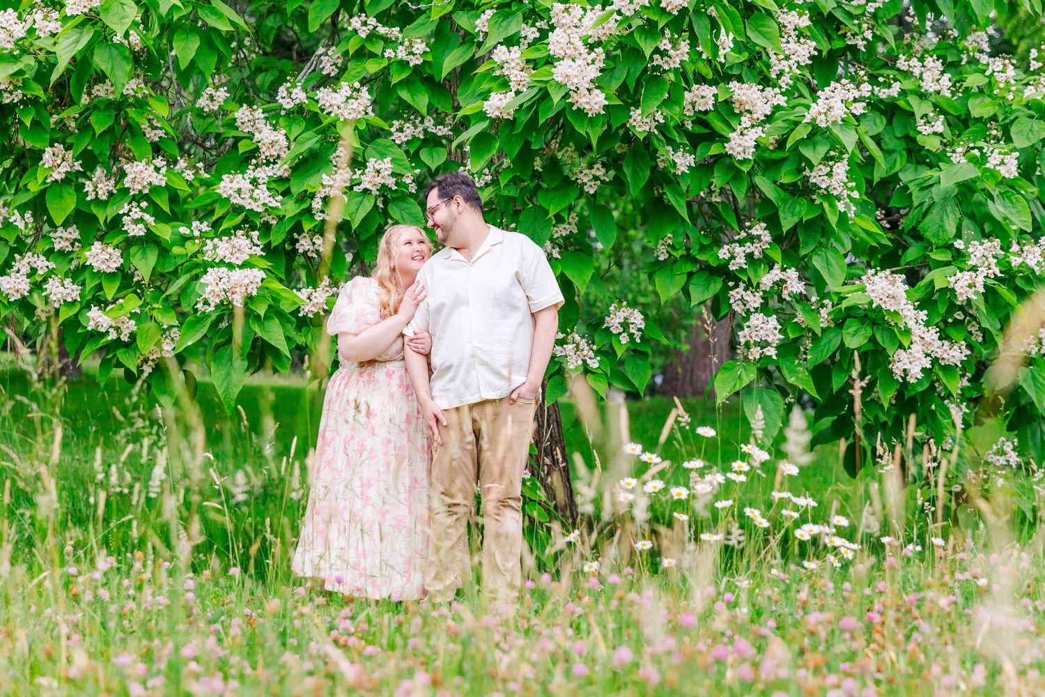 A couple sits affectionately together in a wildflower meadow surrounded by lush green trees on a sunny afternoon.