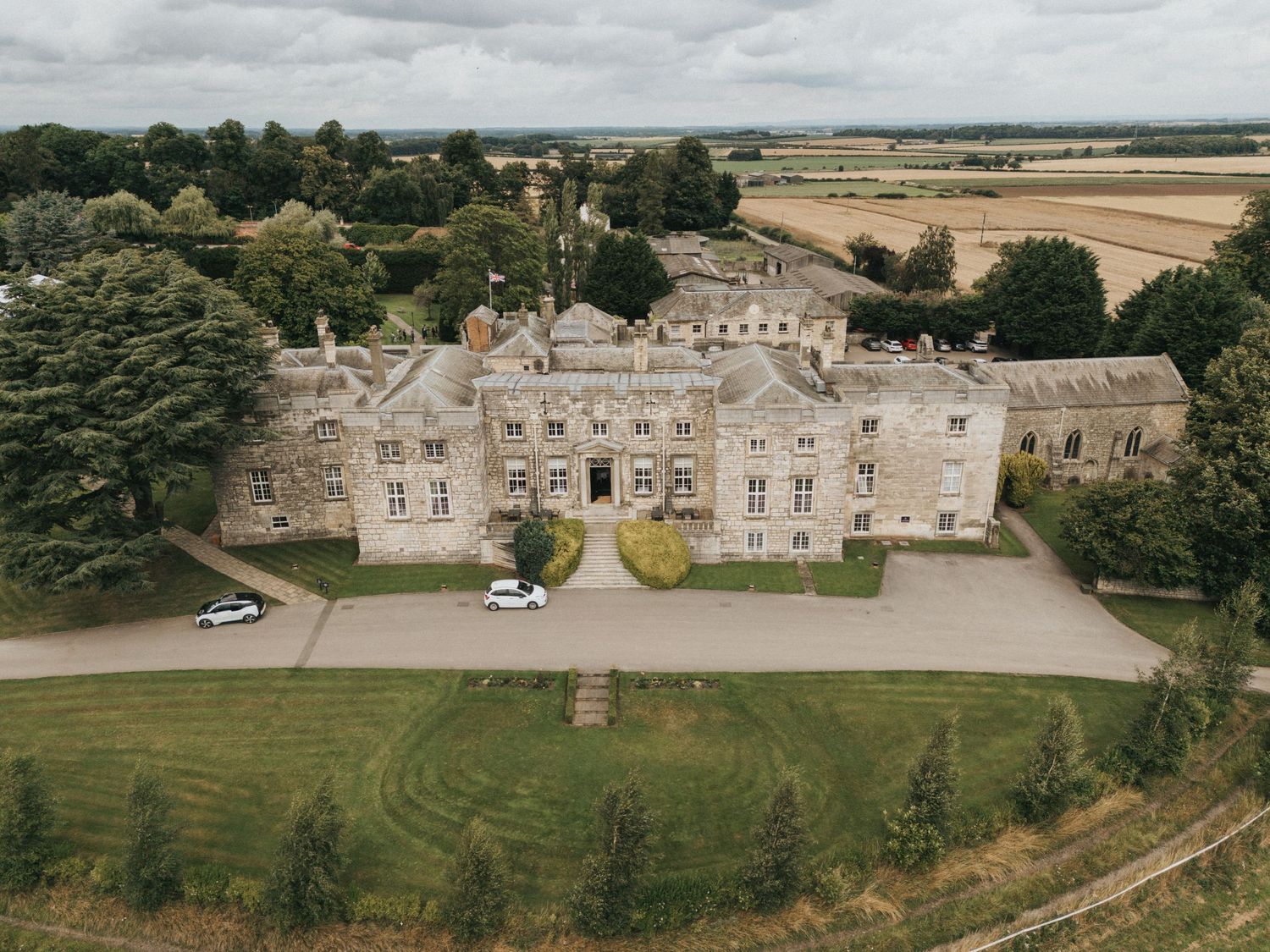 Aerial view of a large stone manor house with manicured lawns and gravel driveway in the English countryside.