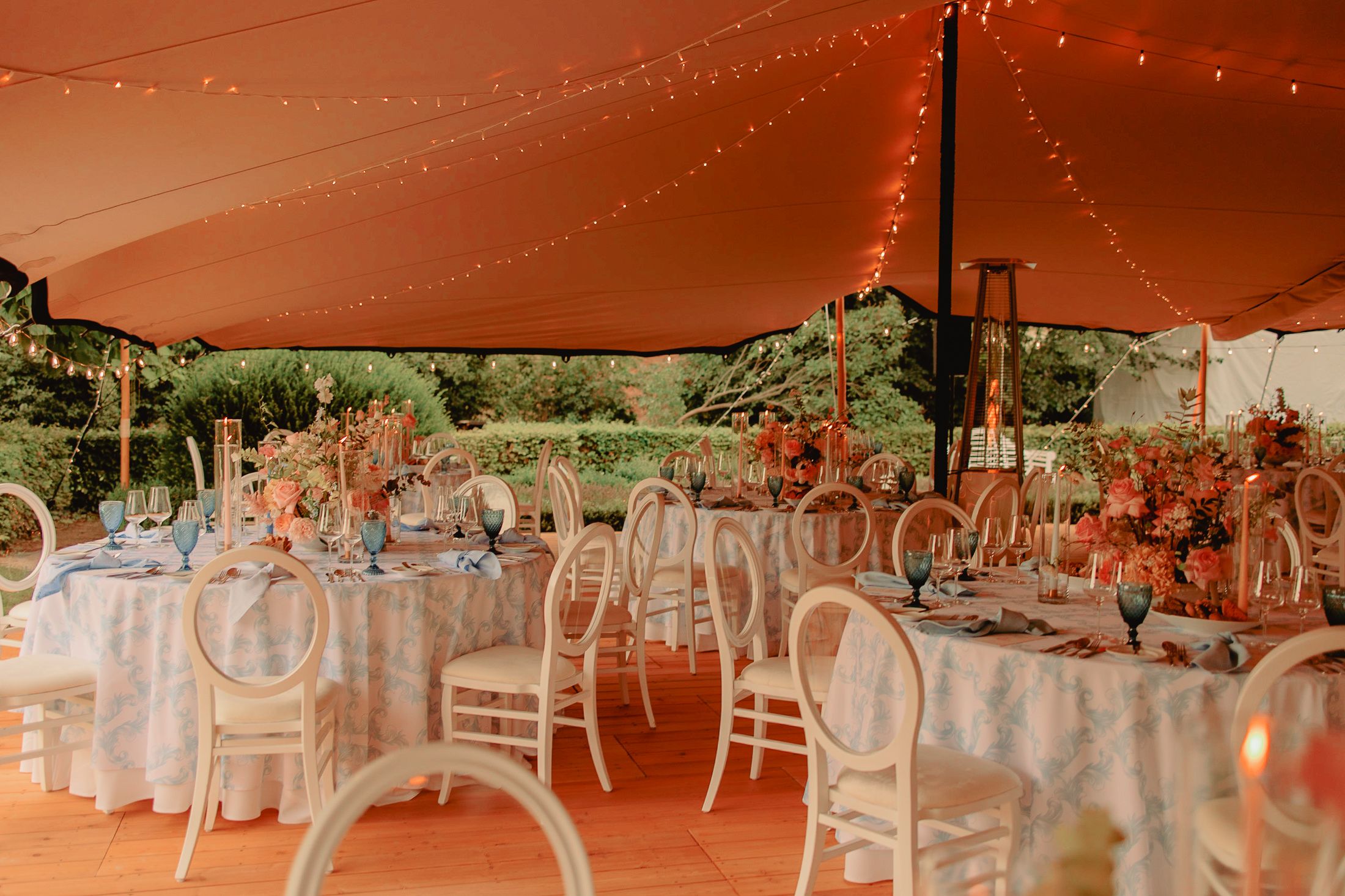 Elegant wedding reception setup under a coral-colored tent with white chairs and floral centerpieces on round tables.