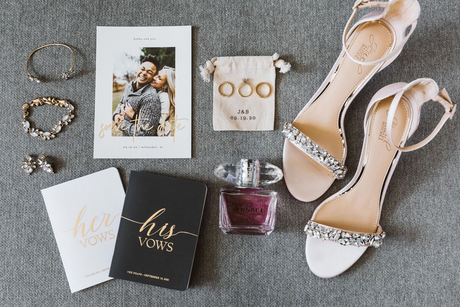 Wedding accessories including rings, shoes, and invitations arranged on grey fabric.