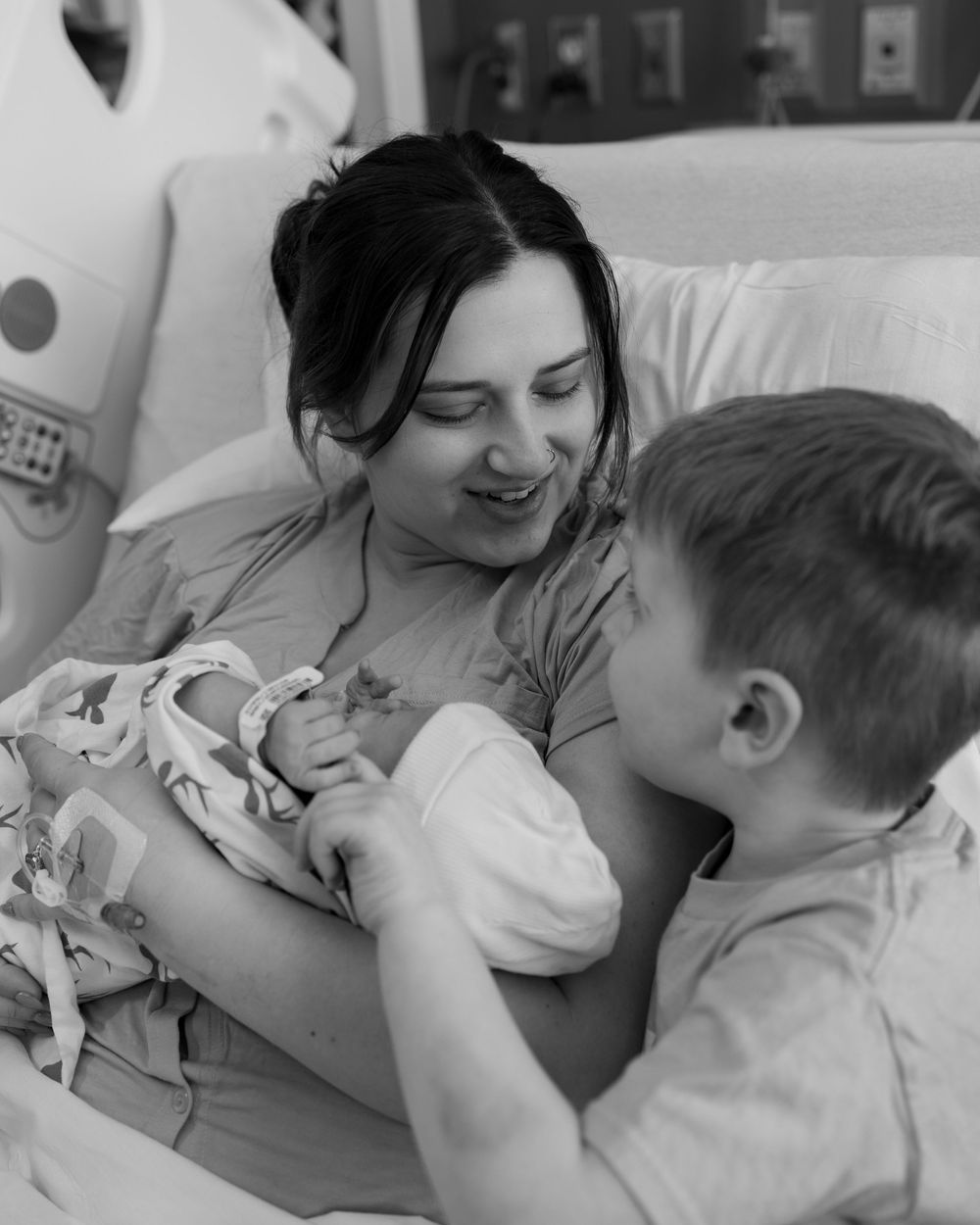 Black and white image showing loving moment between a mother in a hospital bed with her newborn and young child.