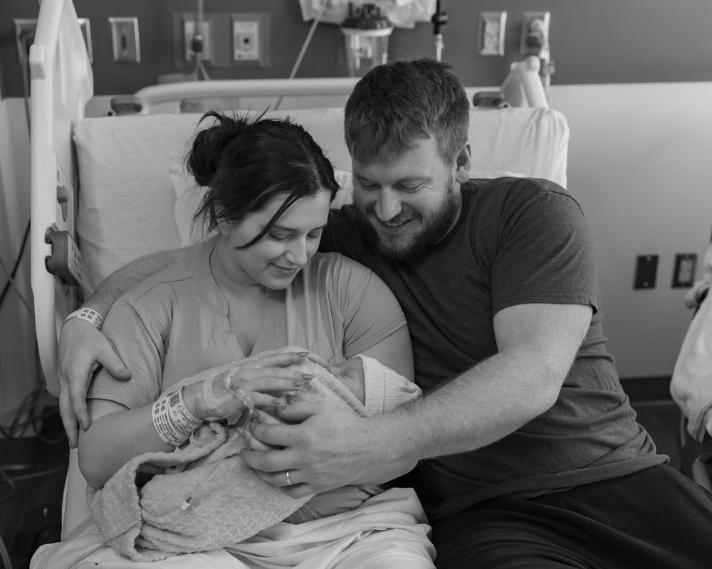 Black and white photo showing a couple tenderly holding and gazing at their newborn baby in a hospital room.