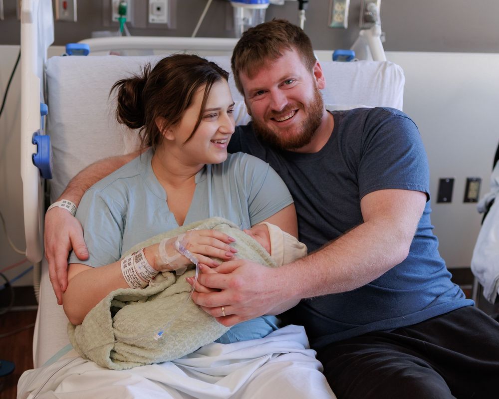 A couple in a hospital room smiles joyfully while holding their newborn baby wrapped in a soft beige blanket.