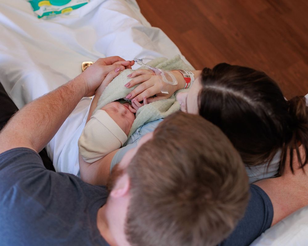 A newborn baby rests peacefully while being cradled by loving arms on a wooden floor.
