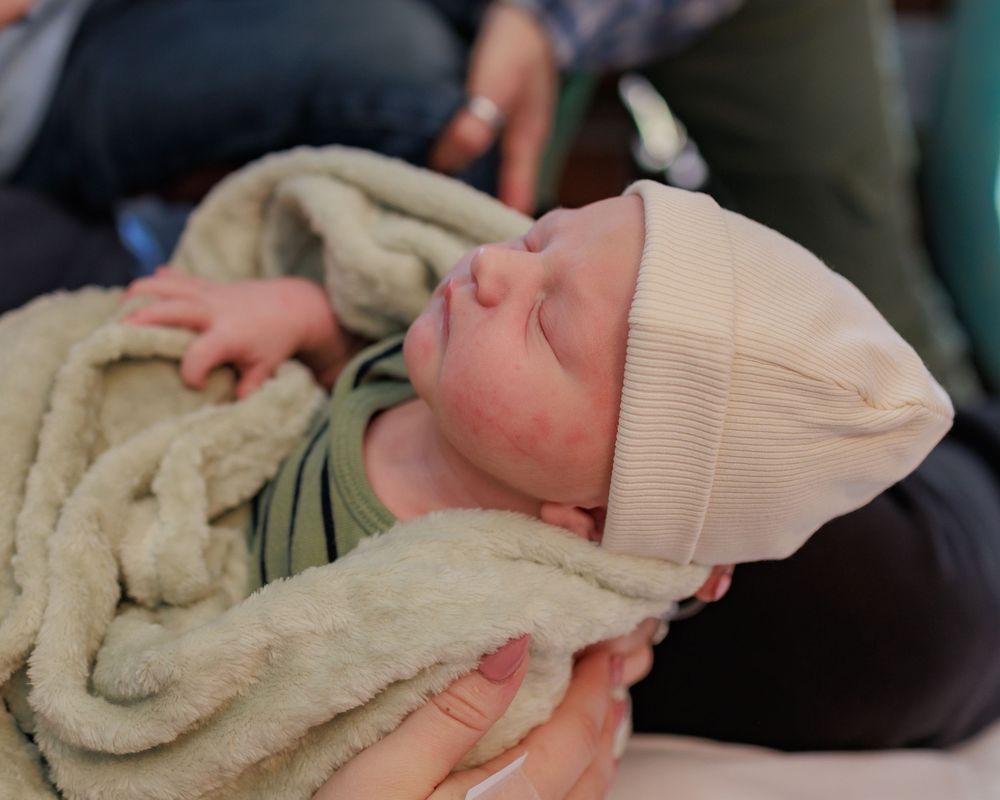 A sleeping infant wearing a cream beanie is wrapped in a soft blanket while being held.