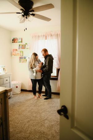 A nursery room view through a doorway shows a couple standing together near a ceiling fan and curtained window.