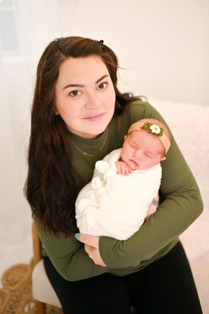 Woman in olive green top holding sleeping newborn wrapped in white blanket in studio setting.