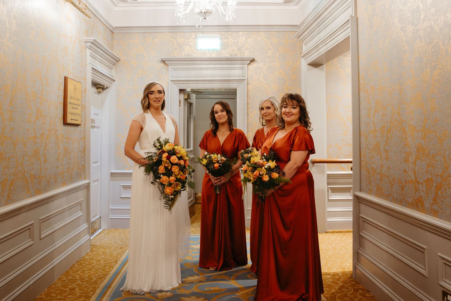 Wedding party members in white and red dresses pose with autumn-colored bouquets in an elegant hallway.
