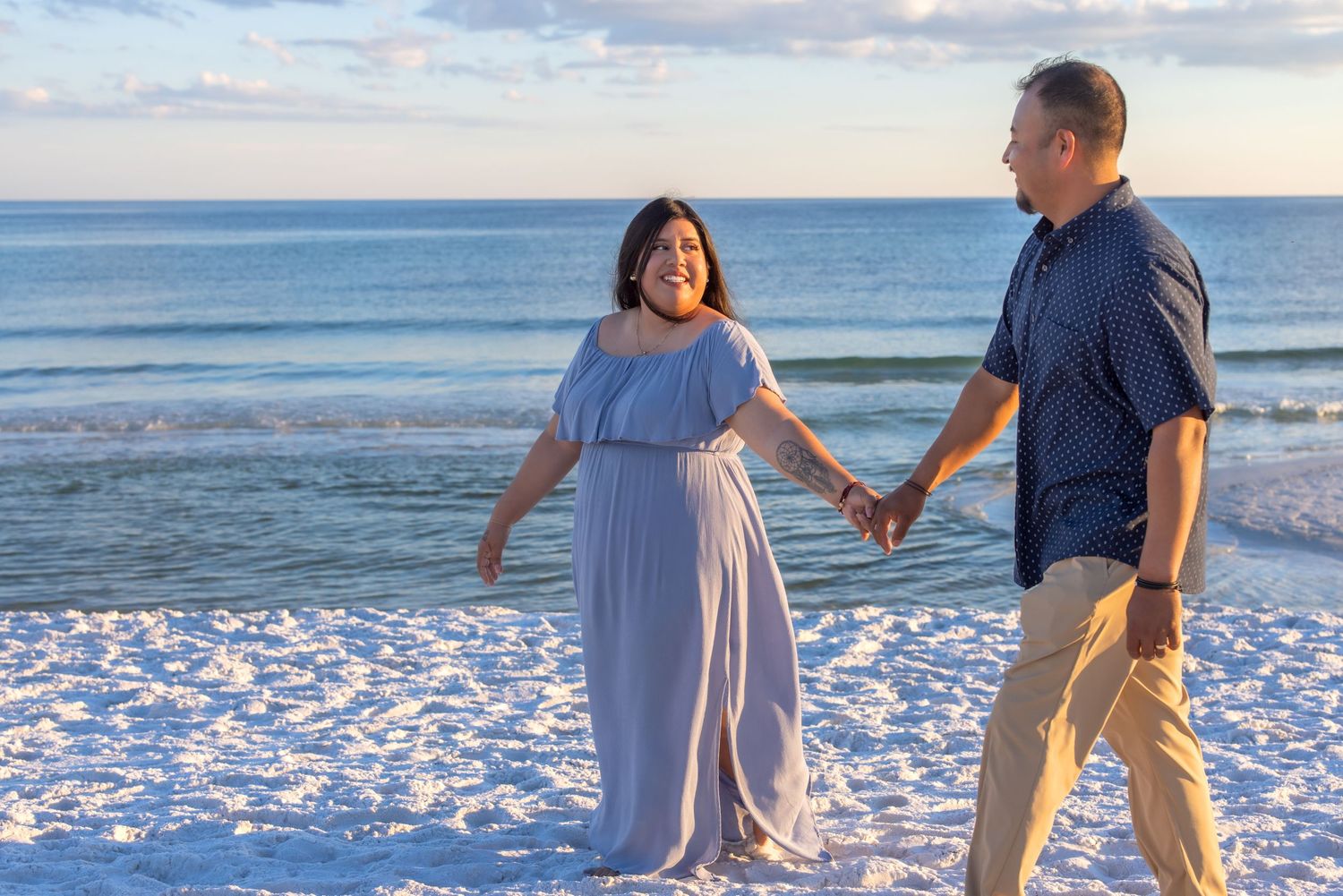 Couple holds hands while standing together on a beach at sunset with ocean waves and cloudy sky in background.