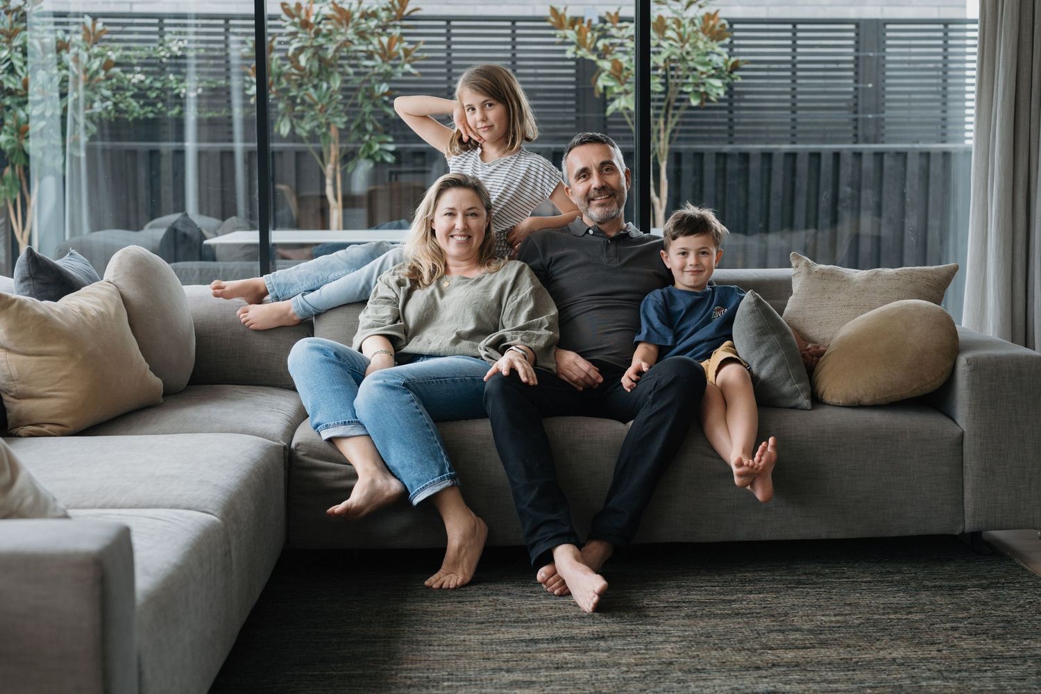 A comfortable family portrait of a mother, father and two children sitting on their home couch smiling to camera.