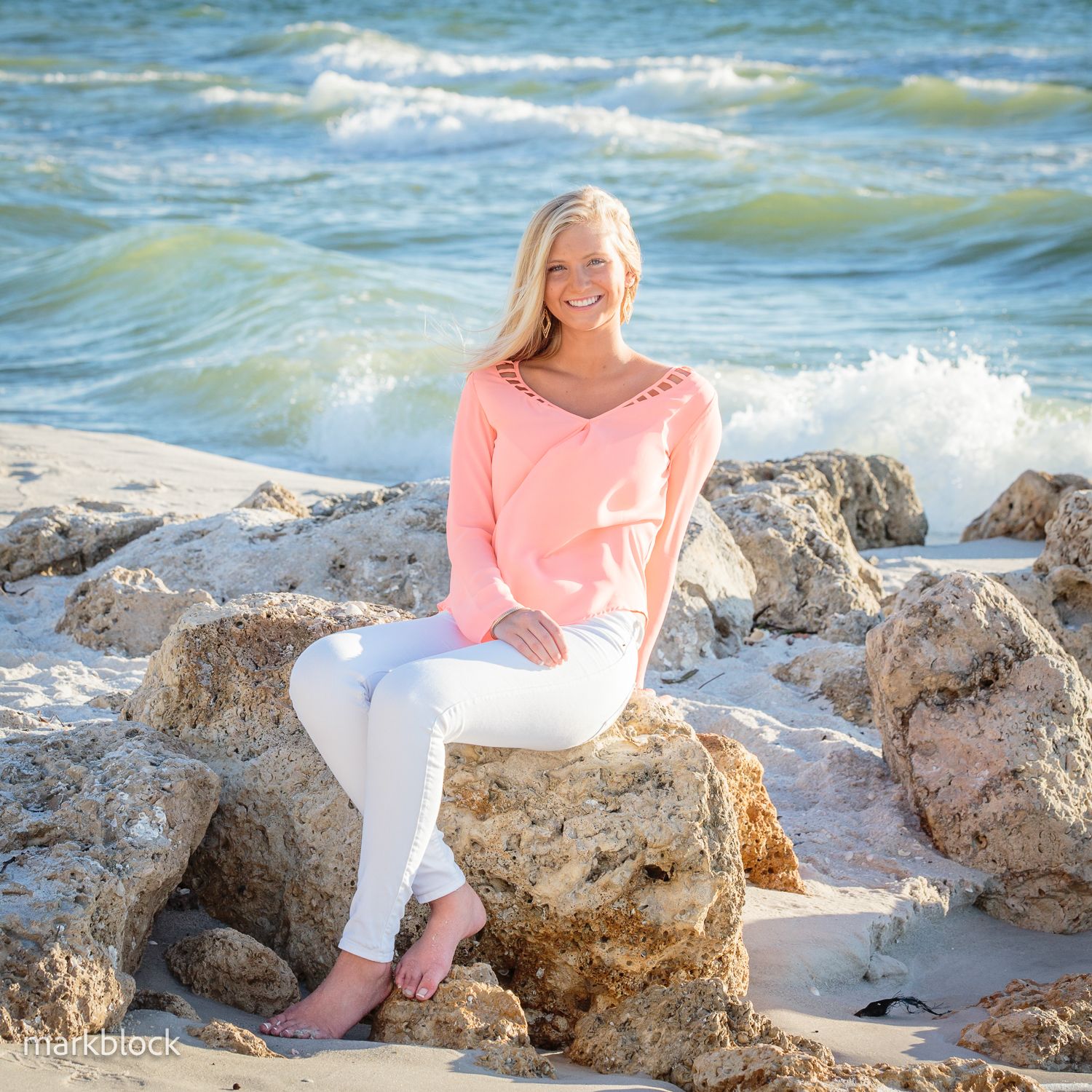 A person in a pink sweater and white pants sits on coastal rocks with ocean waves crashing in the background.