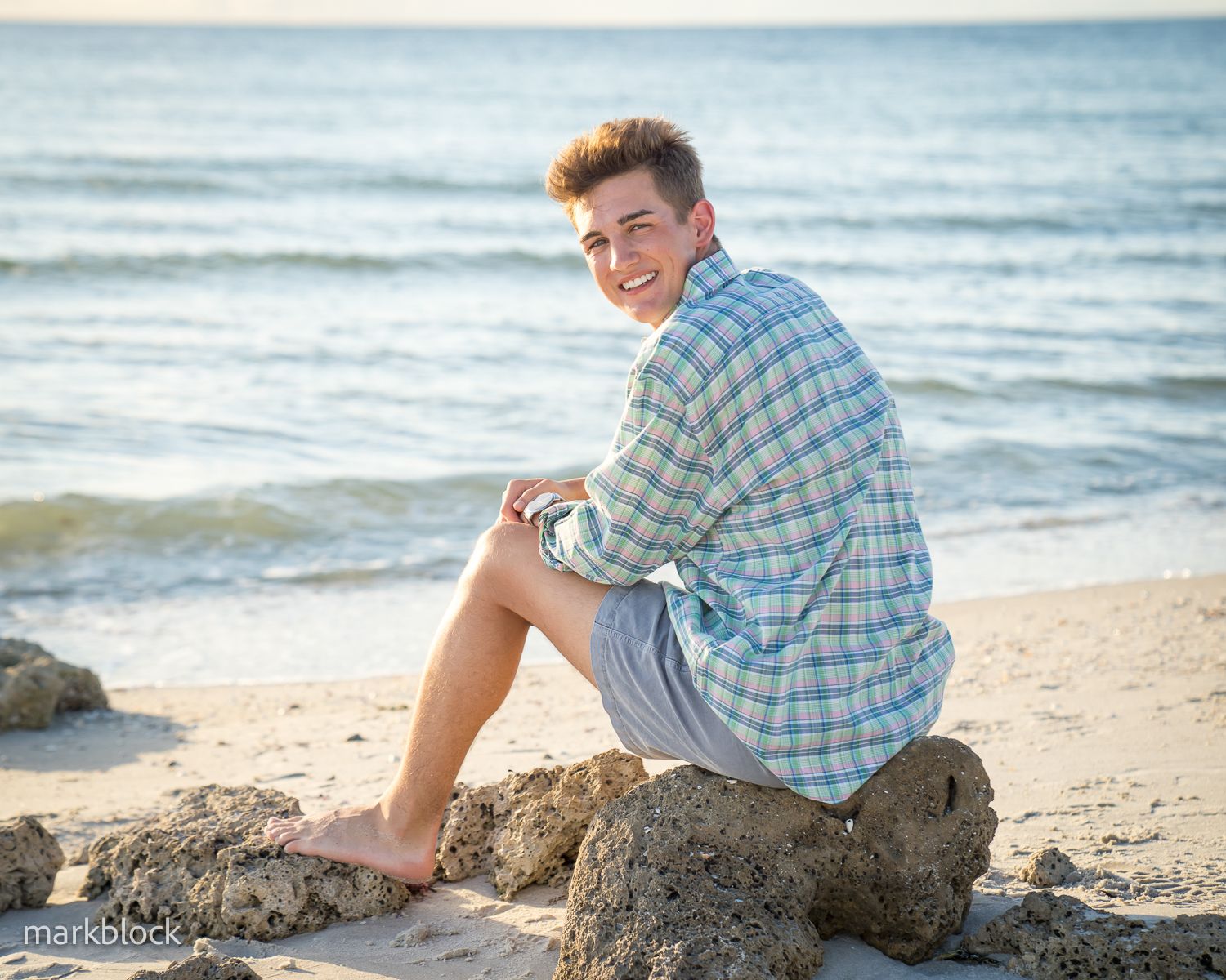 Someone sits barefoot on a rocky beach wearing a plaid shirt as ocean waves roll in during sunset.