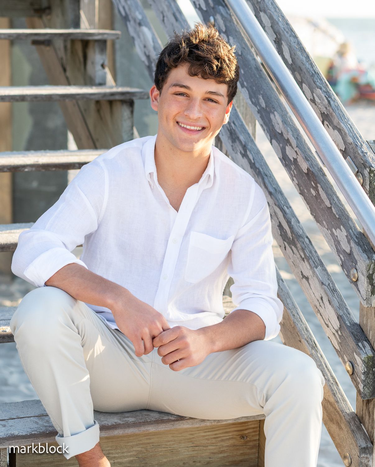 A young person wearing white shirt and beige pants sits casually on wooden stairs outdoors in natural lighting.