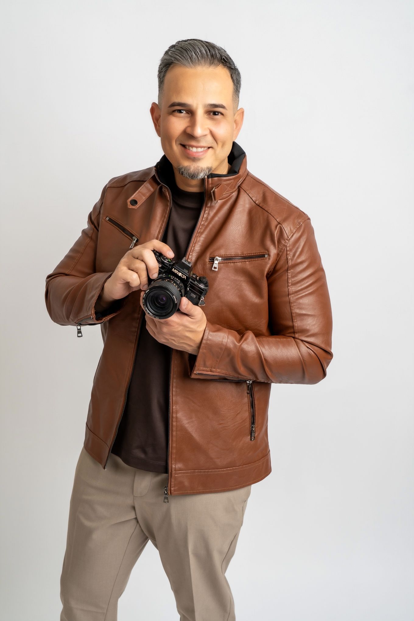 Photographer in brown leather jacket holds professional camera against white studio background.