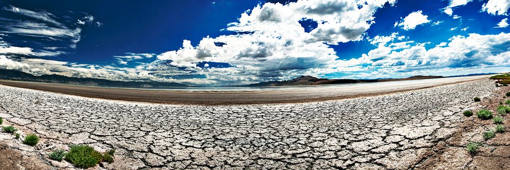 Panoramic view of a cracked dry lake bed with white salt surface stretching to the horizon under dramatic blue skies.