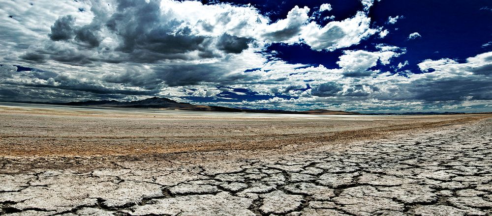 Dramatic panoramic landscape of cracked dry desert ground under stormy clouds and deep blue sky.