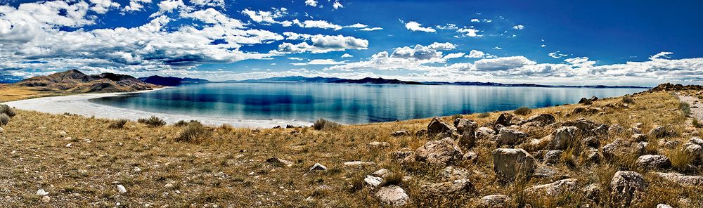 Panoramic view of Lake Tso Moriri in Ladakh with crystal blue waters surrounded by rocky shores and dramatic clouds.