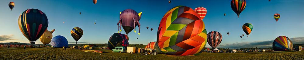 Hot air balloons of various colors lift off at sunrise during a balloon festival against a panoramic sky.