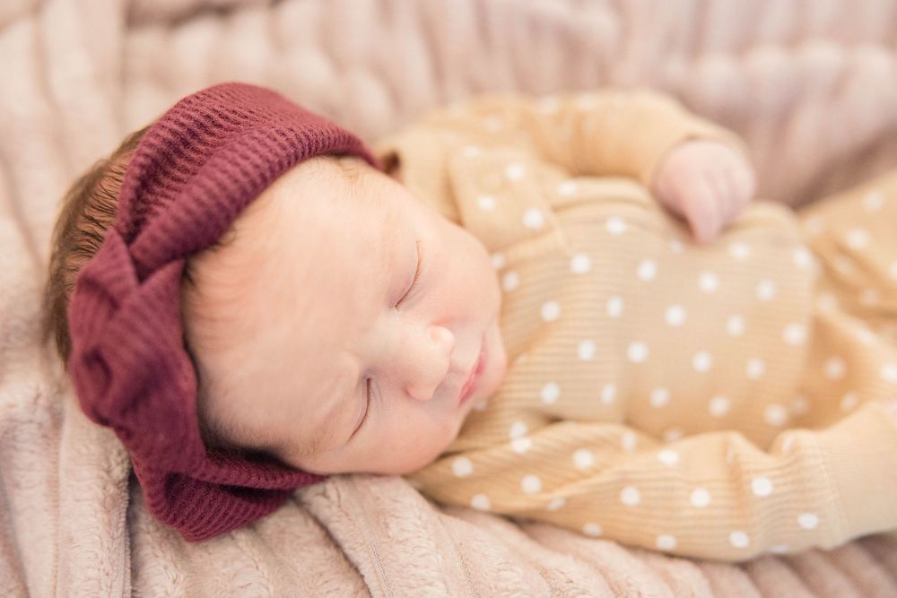 Sleeping baby wearing burgundy knit beanie and polka dot sleeper rests peacefully on soft blanket.
