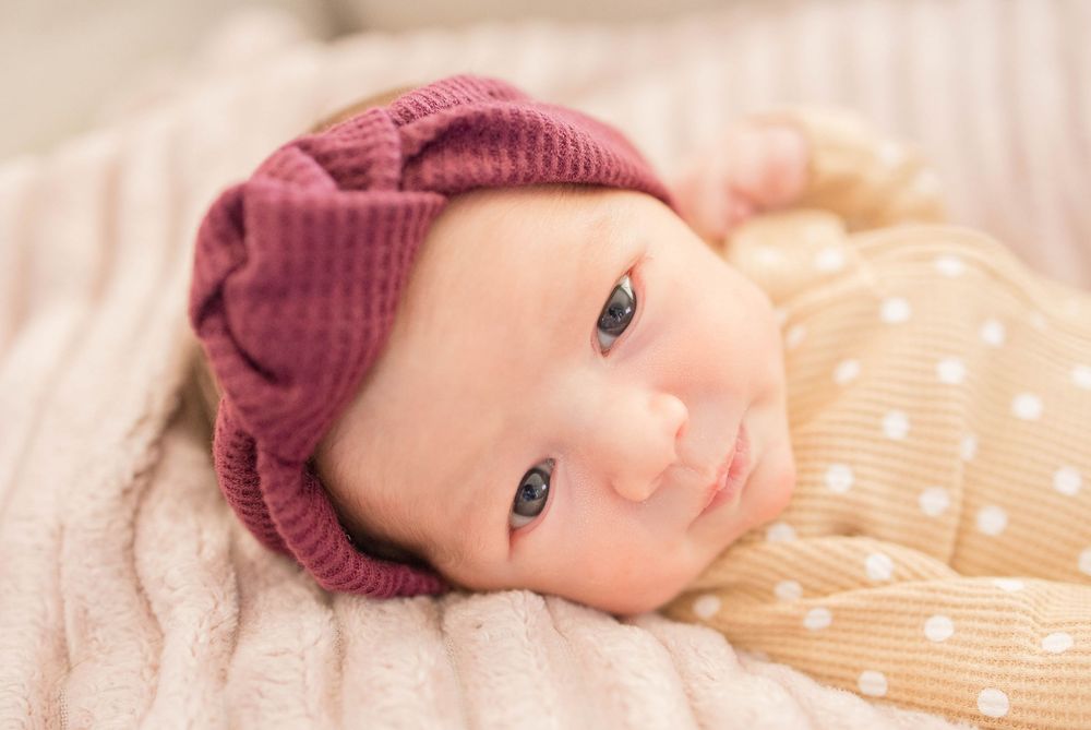 A baby wearing a burgundy knit hat lies on a polka dot blanket looking at the camera.