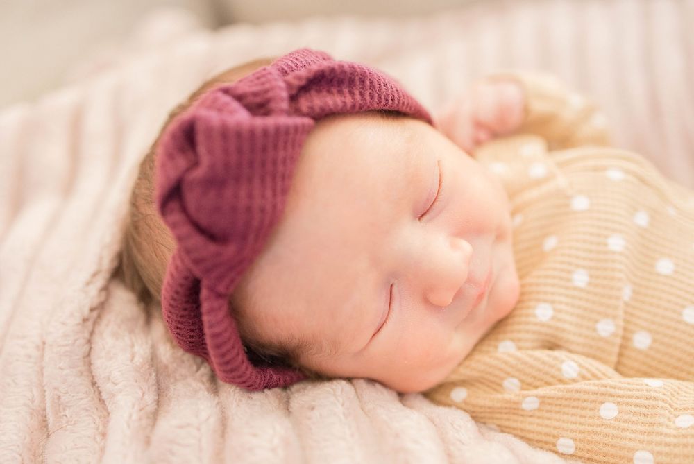 Sleeping baby wearing a burgundy knit hat rests on polka dot blanket in soft natural lighting.