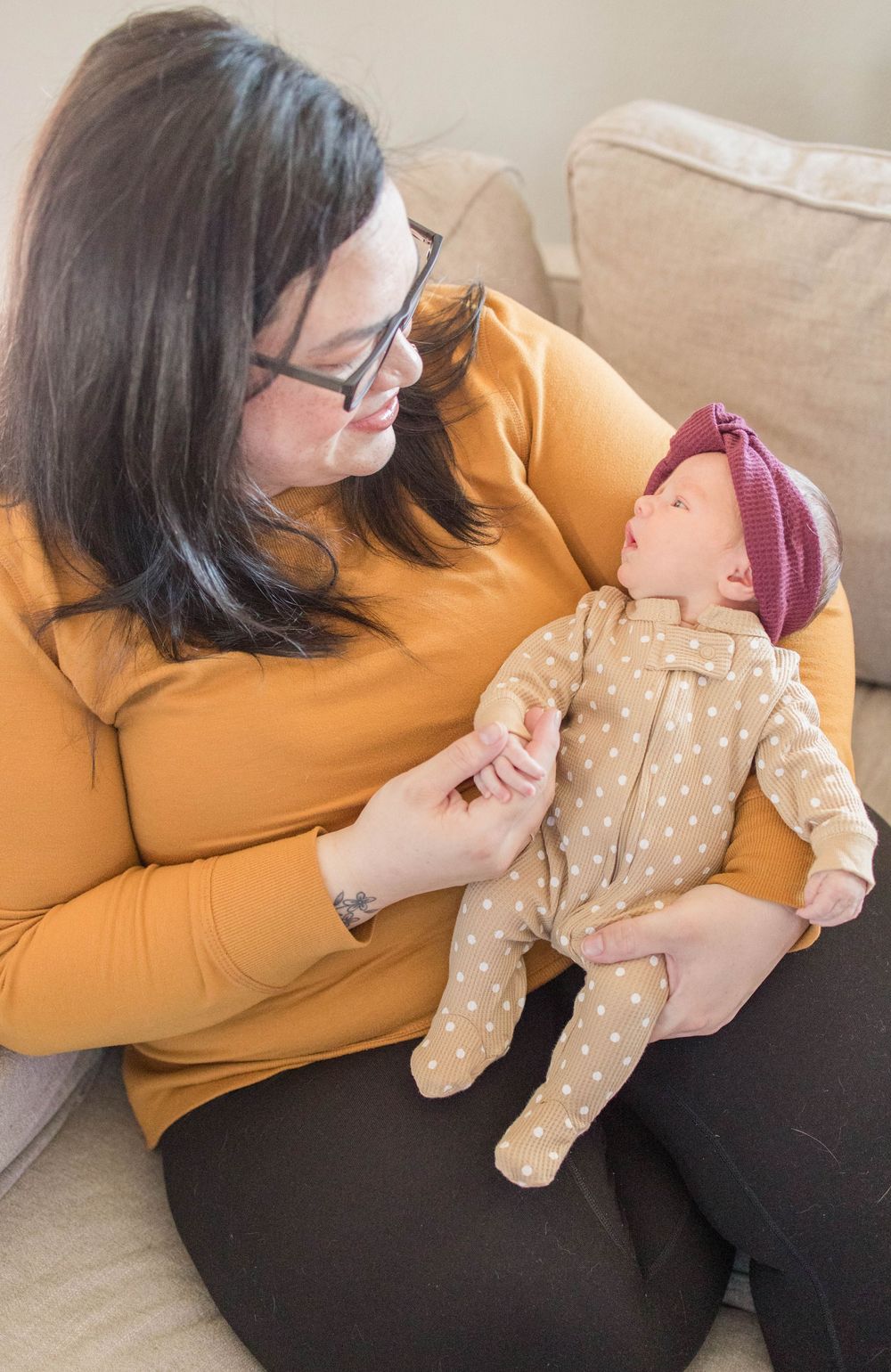 Person wearing an orange sweater gently holds a baby dressed in polka dot pajamas and burgundy hat on a beige couch.