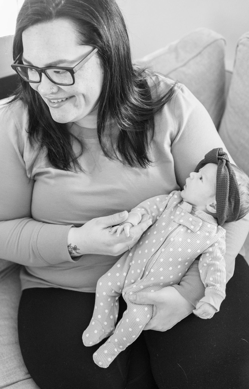 Person in glasses holding a polka dot onesie-clad baby doll while sitting on a couch in black and white.
