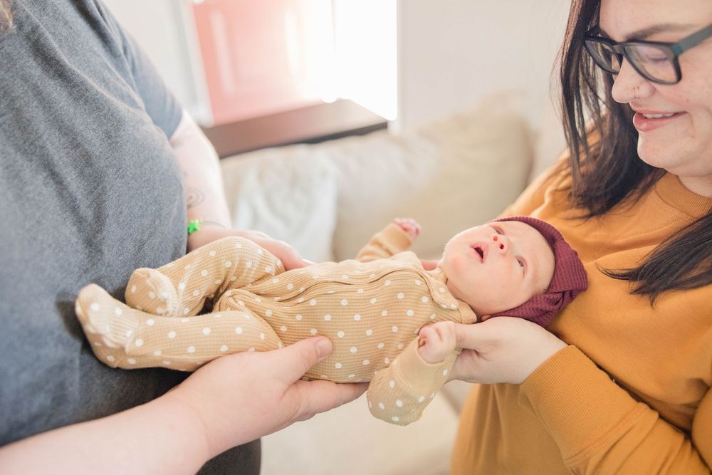 Baby in beige onesie being gently held by adults with caring hands in soft natural lighting.