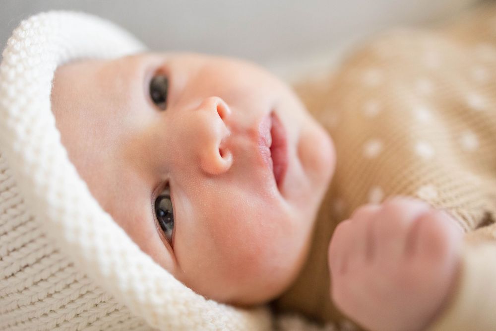 Infant wearing white knit hat gazes peacefully upward while lying on soft beige blanket.