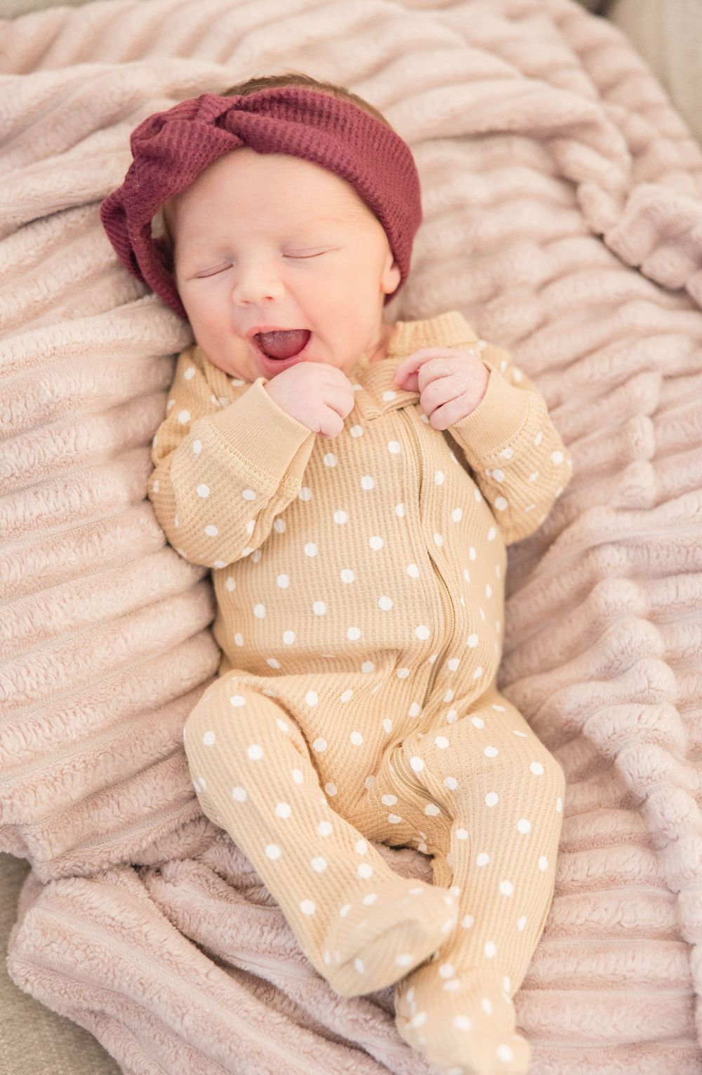 A baby wearing a burgundy beanie and polka dot onesie yawns while lying on a plush blanket.