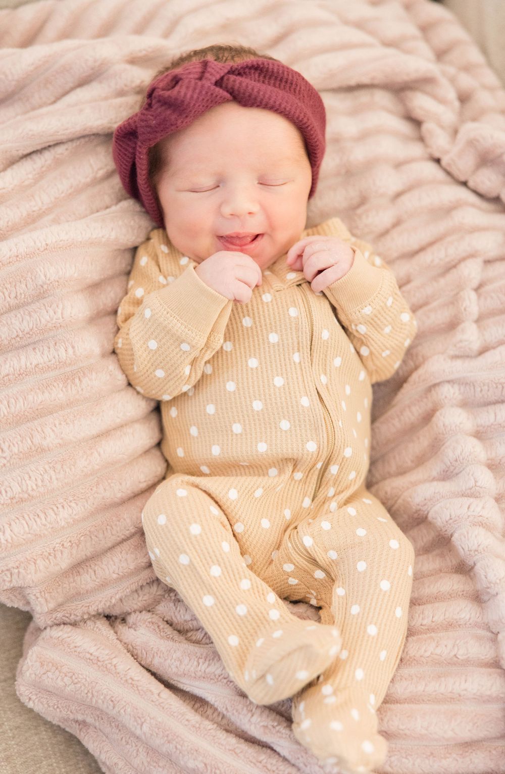 Sleeping newborn wearing a burgundy knit hat and beige polka dot onesie rests on soft pink blanket.