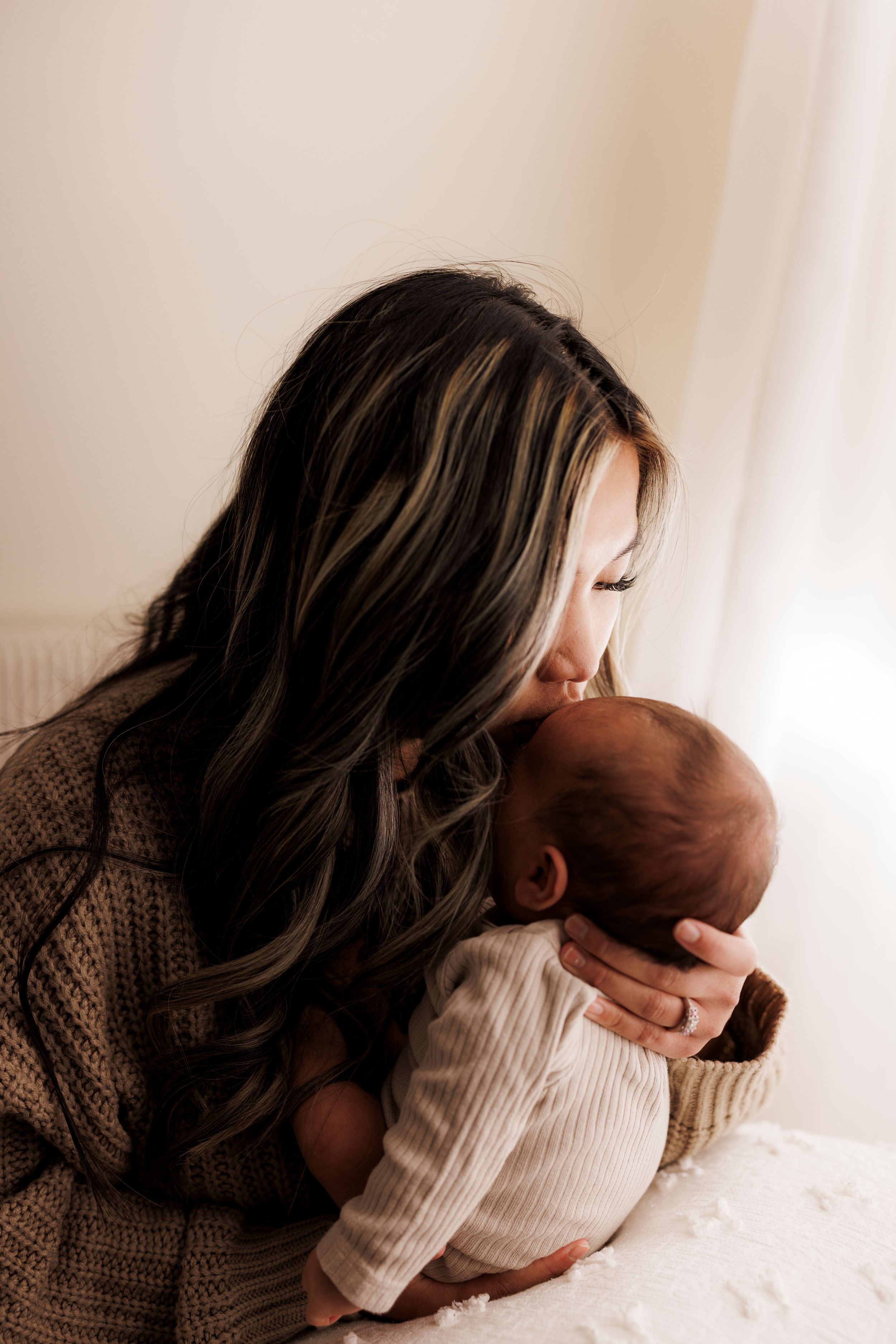 A tender moment between a mother in a brown sweater and her newborn baby near a window with soft natural lighting.