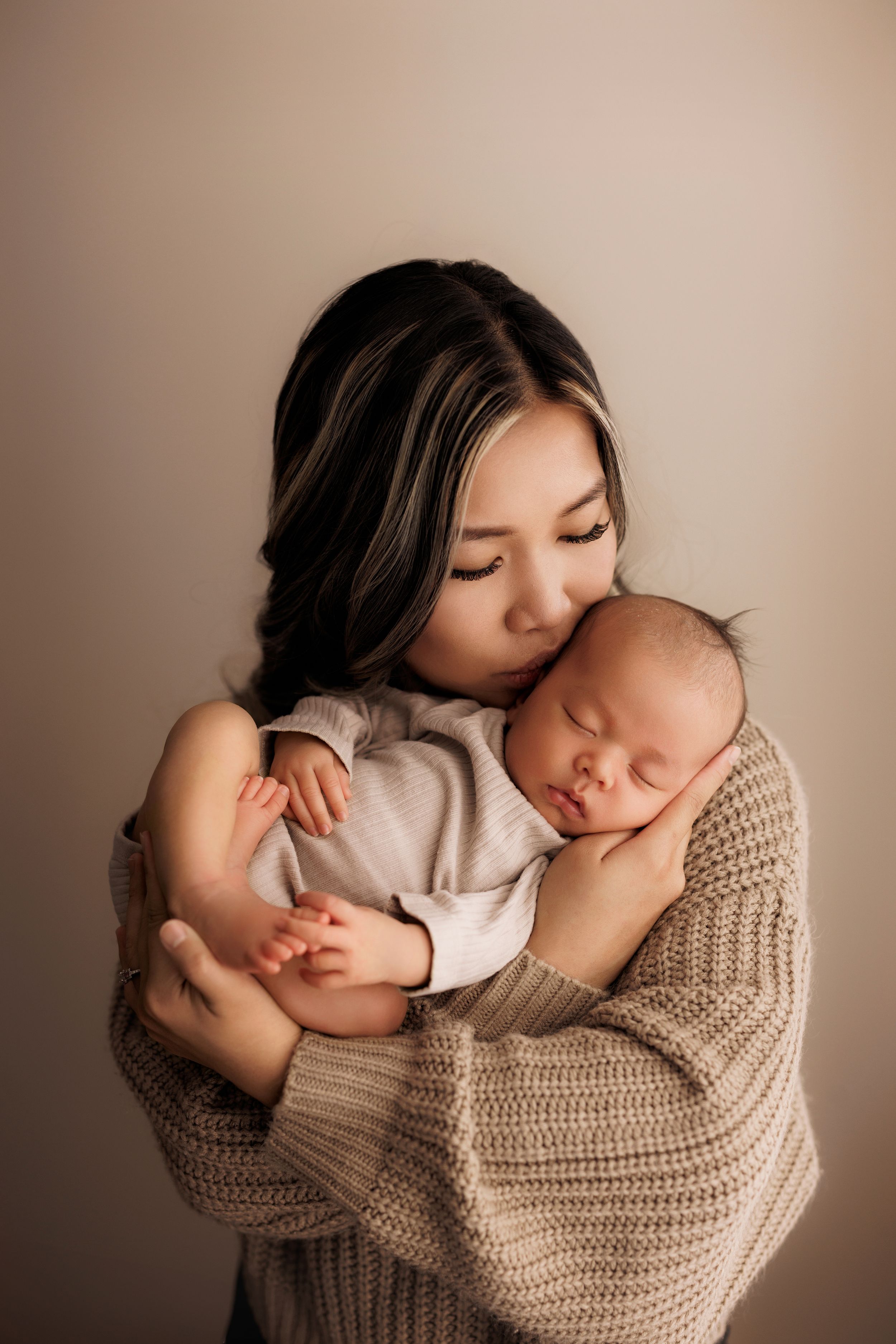 A tender moment between a woman in a knit sweater and sleeping infant captured in warm neutral tones.