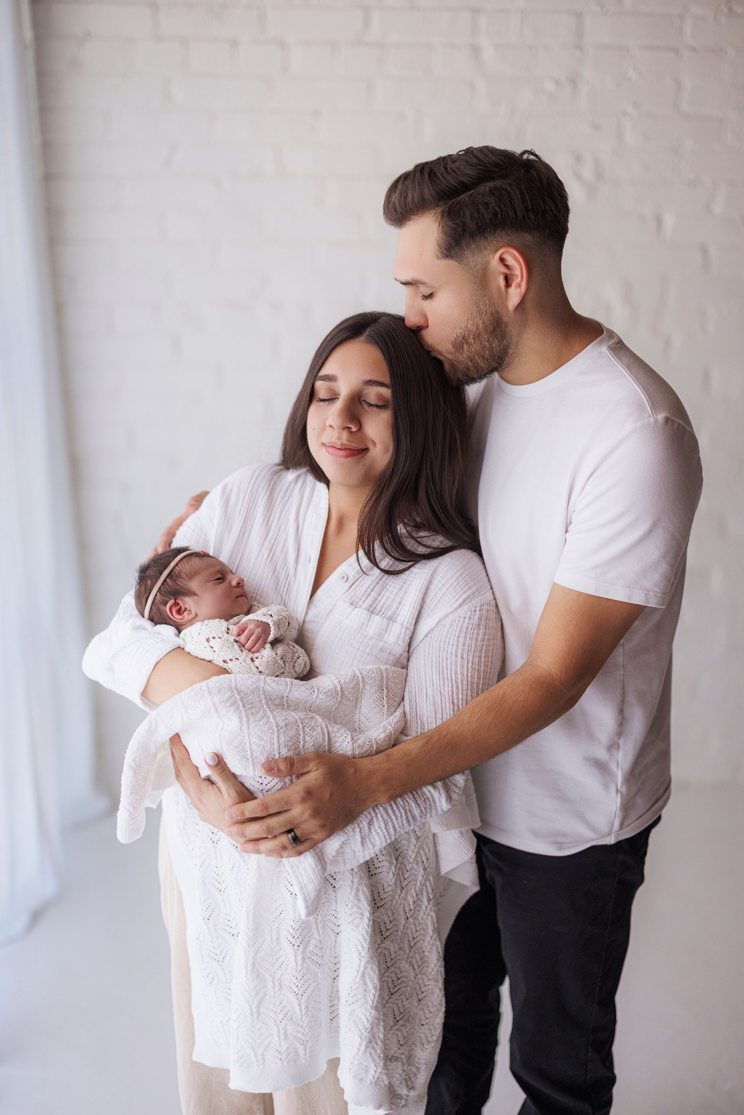 Family in white outfits holding newborn baby during intimate indoor photo session with natural lighting.