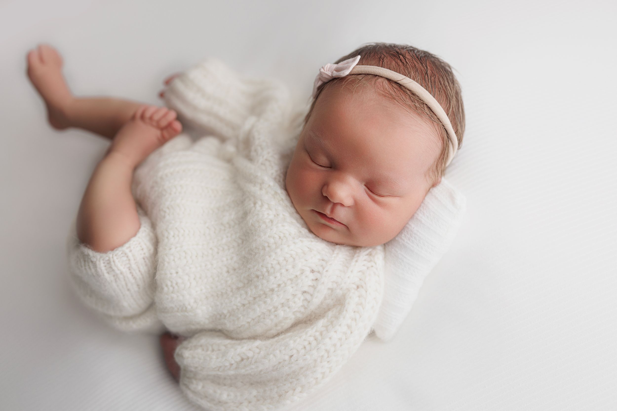 Newborn baby wearing white knit sweater sleeps peacefully on soft white blanket during professional photo session.
