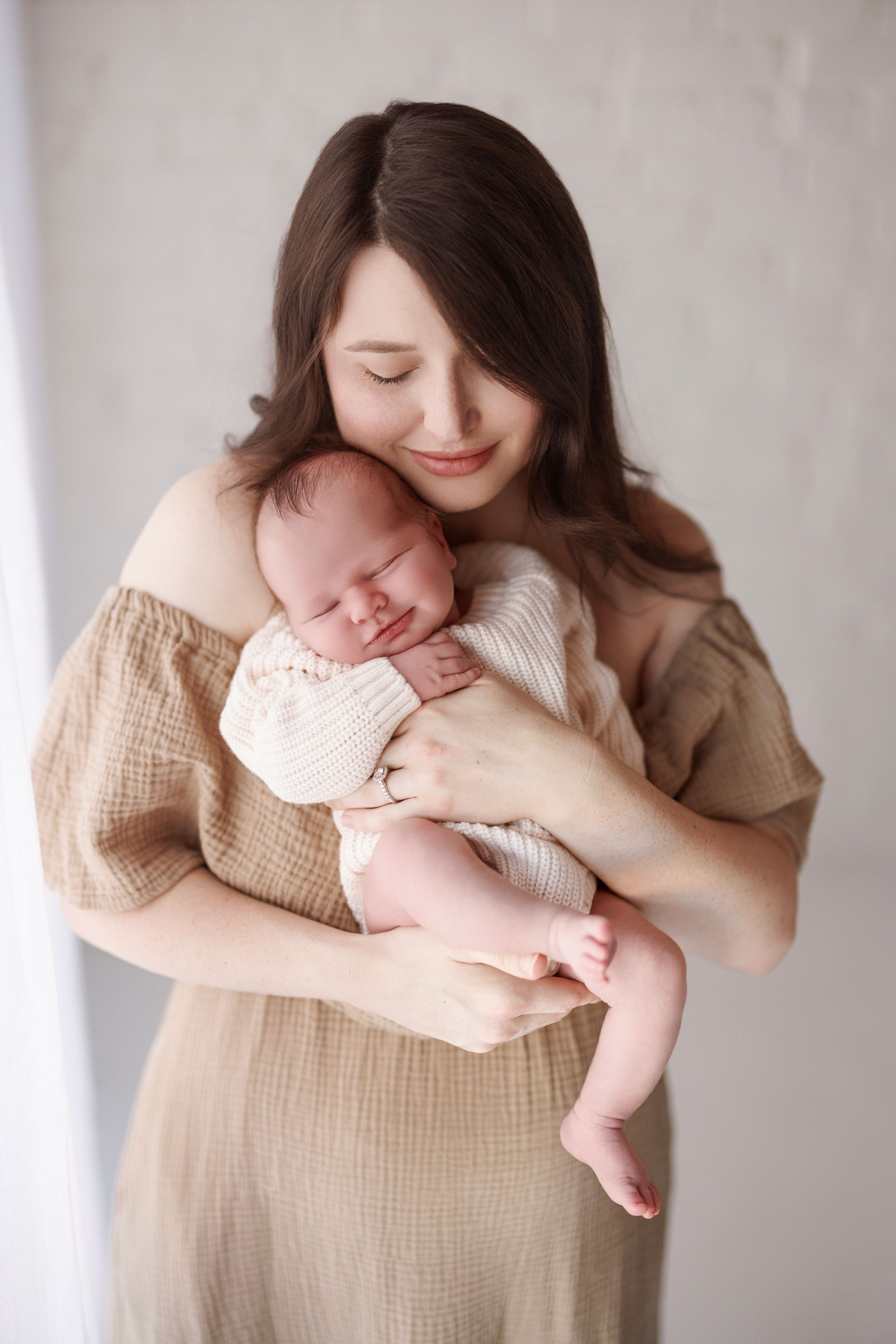 A tender newborn photography session shows a parent in beige muslin fabric cradling a sleeping infant against a light backdrop.
