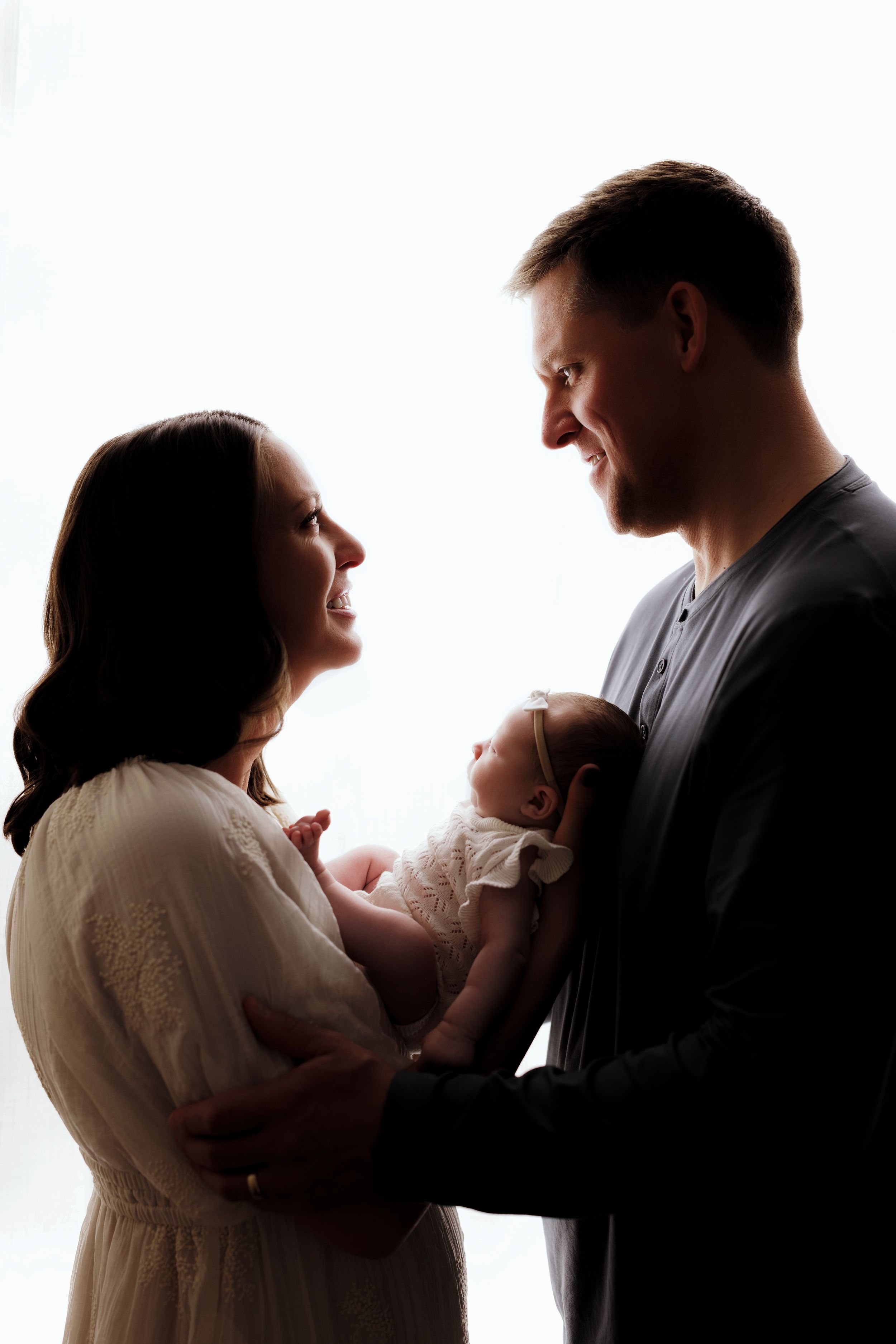 Silhouetted in white light, parents lovingly hold their newborn baby while sharing a tender moment together.