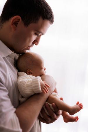 A tender moment showing a parent in a white shirt cradling a small baby wearing a cream sweater against a bright background.