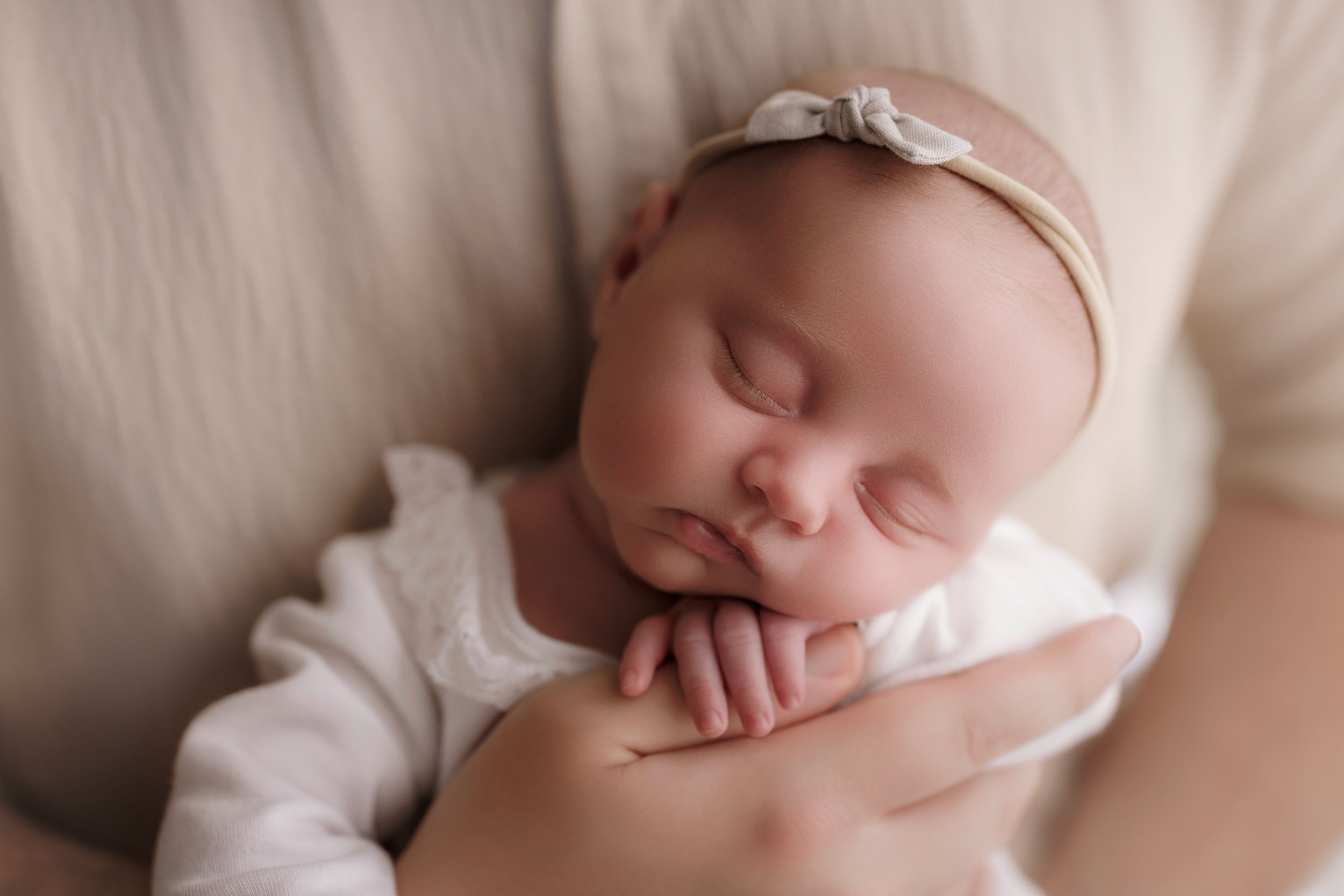 Sleeping newborn baby wearing white sweater and bow headband rests peacefully against soft beige fabric.