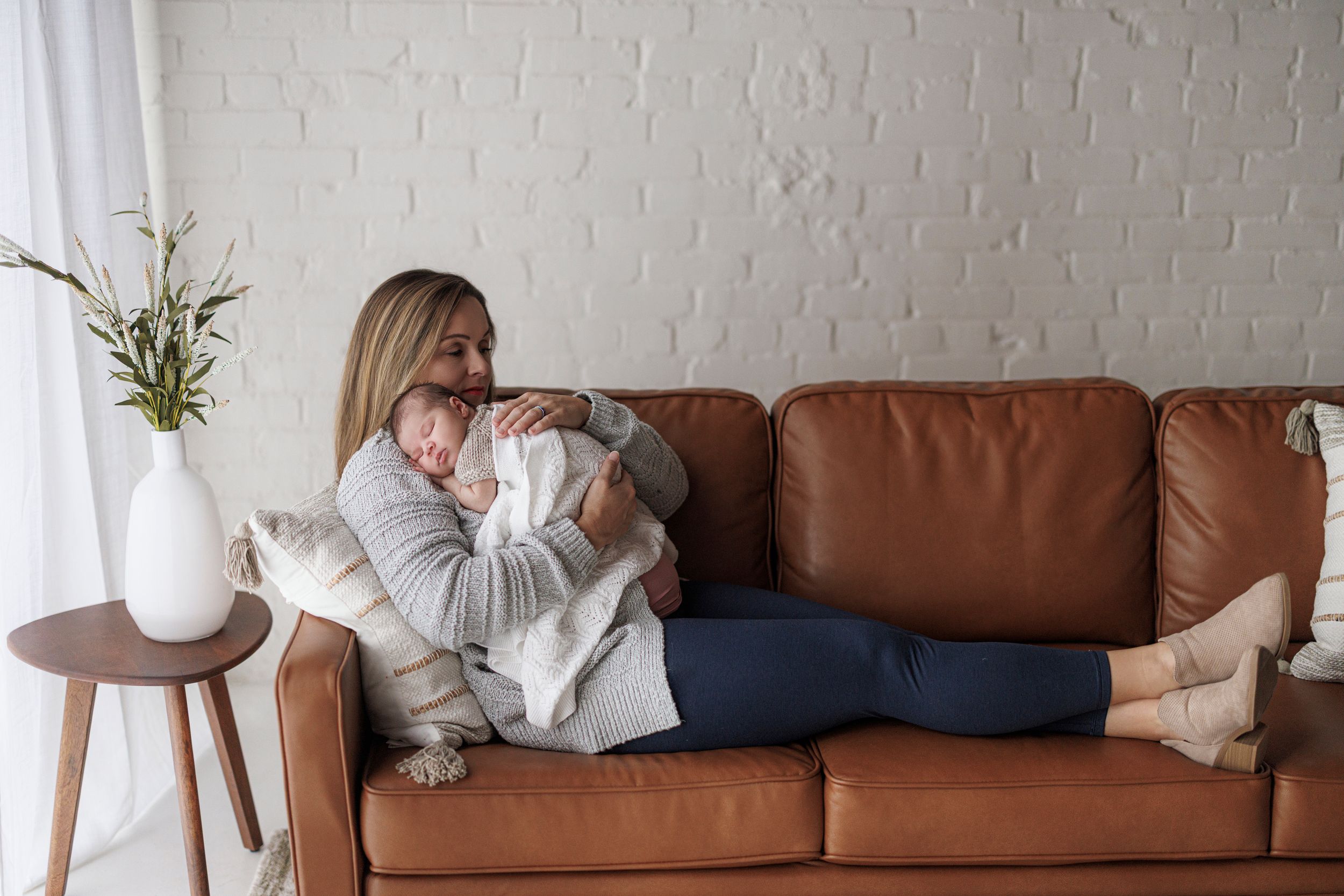 A brown leather sofa sits against a white brick wall with hanging plants and decorative throw pillows.