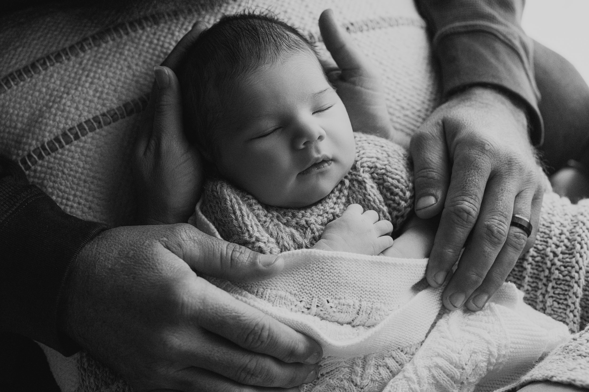 A sleeping baby rests in knitted clothing while being cradled by gentle hands in a black and white photograph.