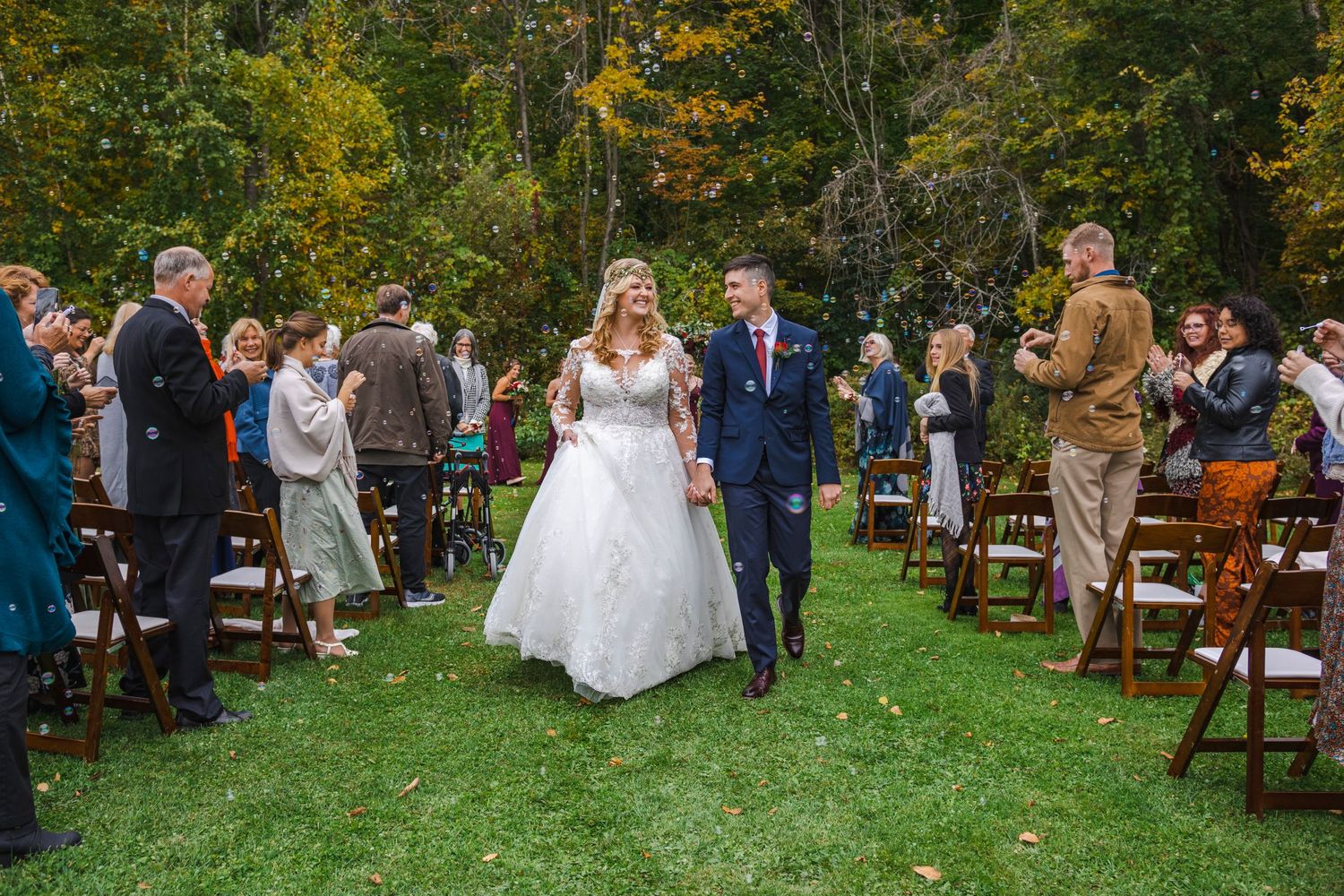 Wedding ceremony outdoors with guests seated on wooden chairs among autumn trees.