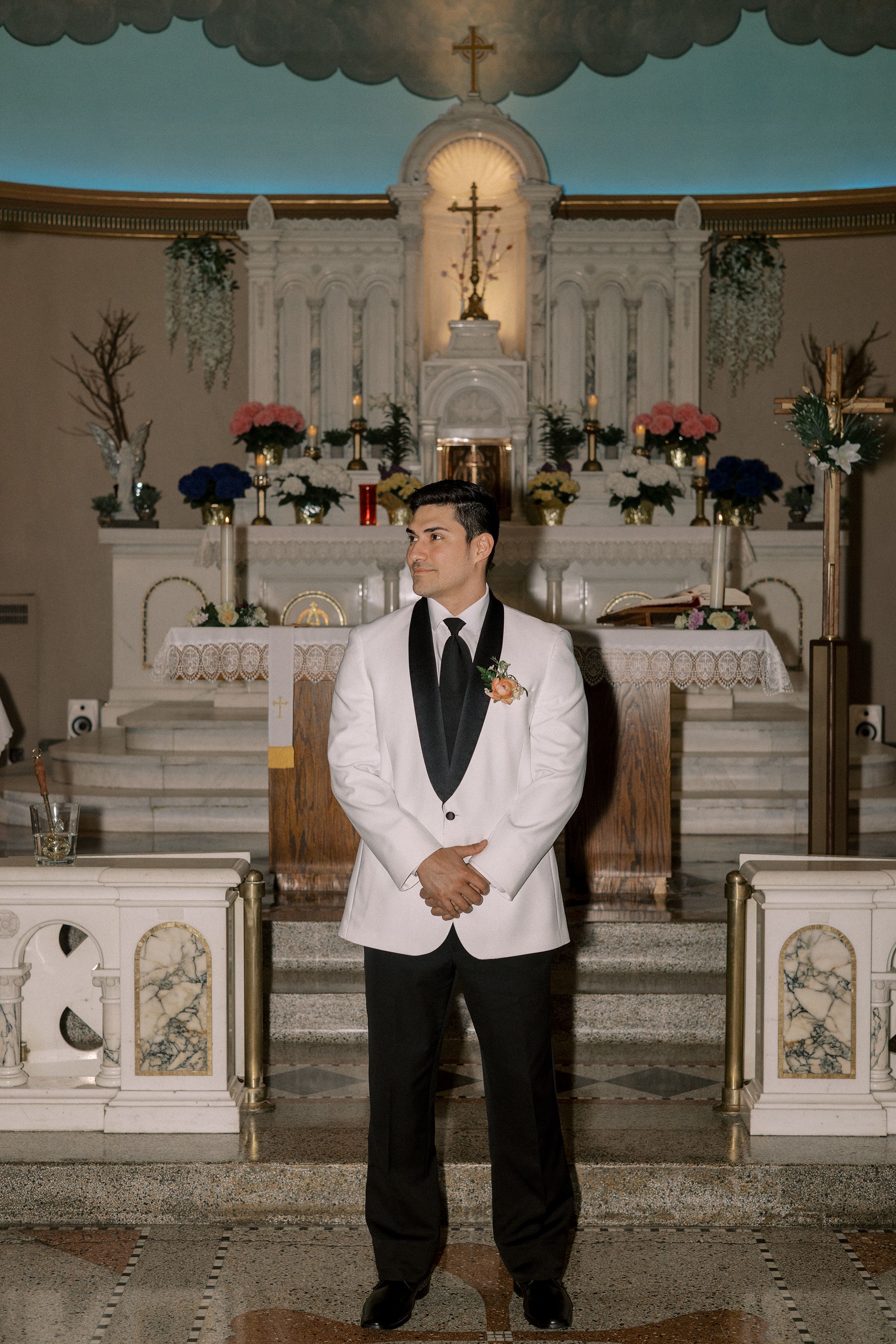Groom in white tuxedo jacket stands at church altar decorated with floral arrangements and crosses.