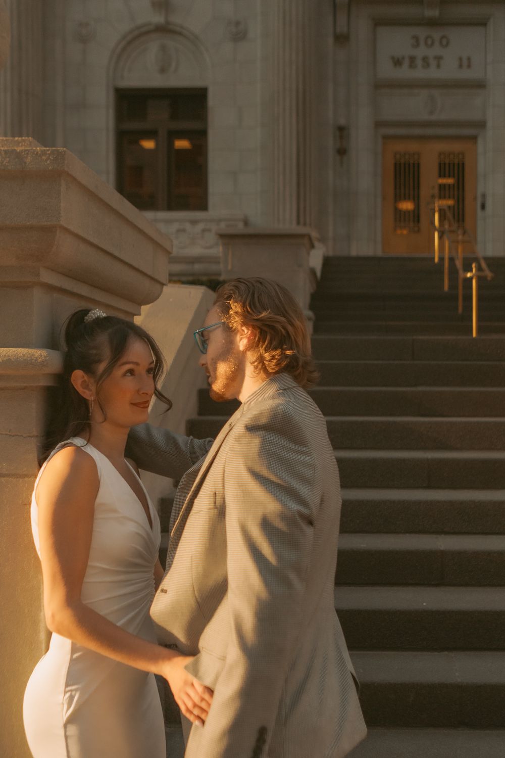 A couple in wedding attire shares an intimate moment on stone steps at sunset in front of an illuminated building entrance.