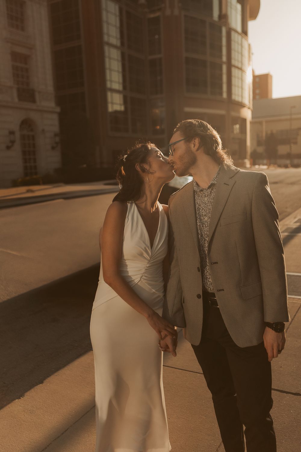 A couple wearing formal attire share a romantic moment on a sunlit city street at golden hour.