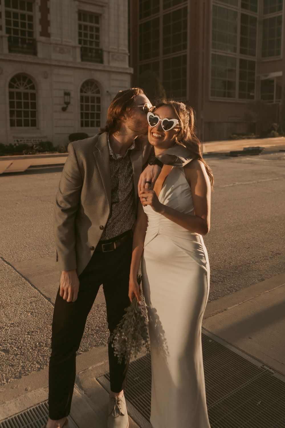 A stylish couple in formal wear share an intimate moment on a sunny urban street.