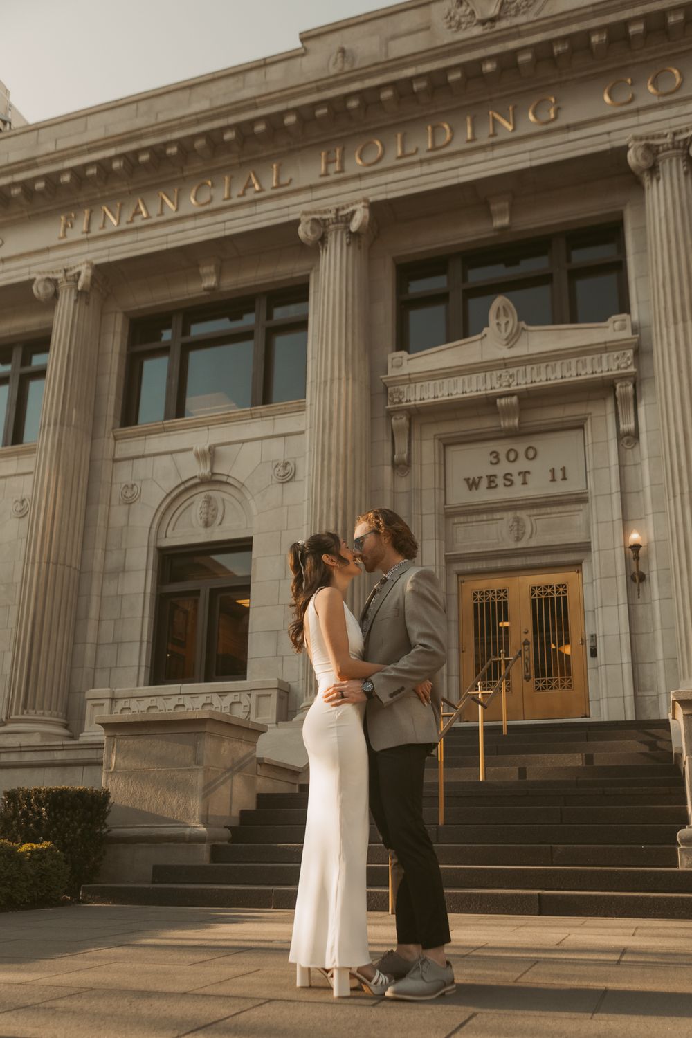A couple stands embracing on the steps of a grand Financial Holding Co. building with classical columns during golden hour.