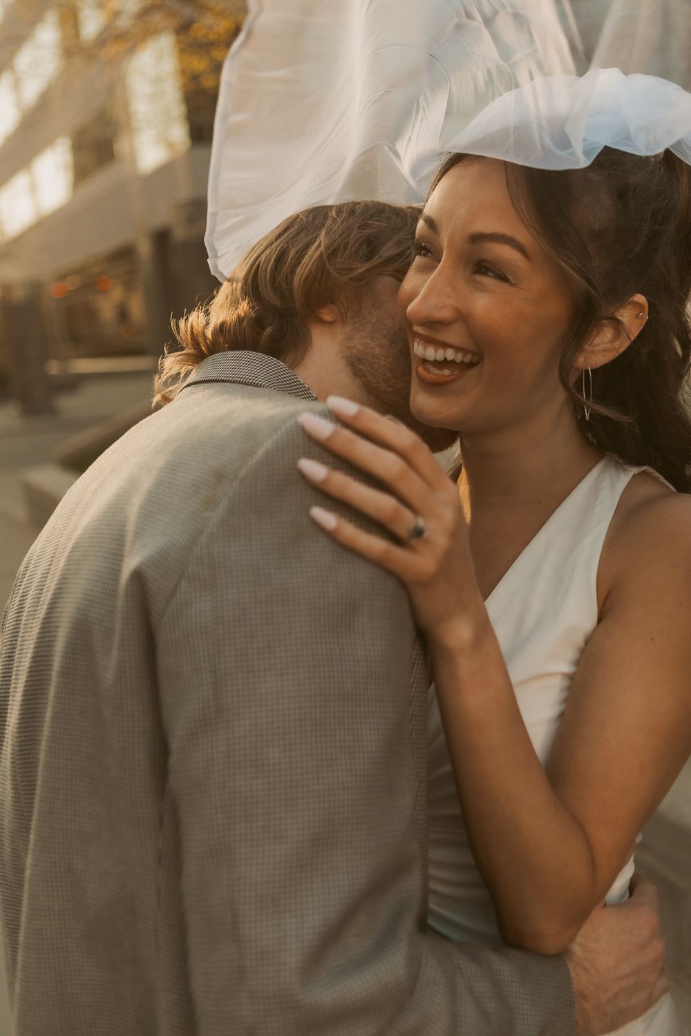 A couple shares an intimate embrace in warm sunlight, with one wearing a white wedding dress and veil.